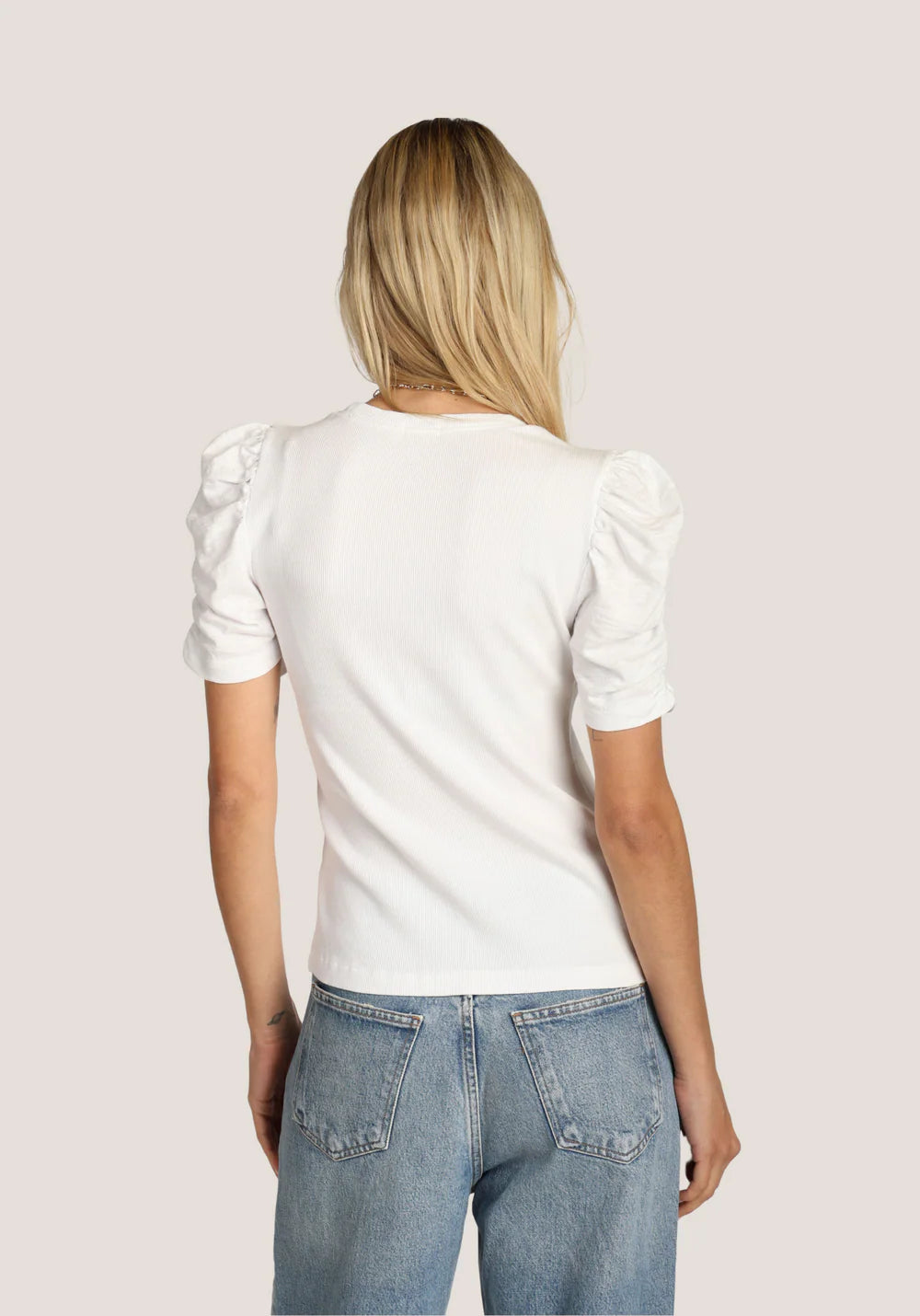 Back view of a woman wearing a white blouse with puff sleeves and blue jeans on a plain background
