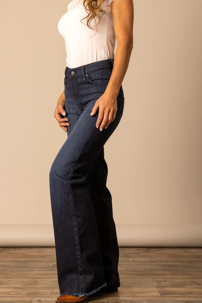 Woman wearing high waisted and dark washed Kimes Olivia flared jeans and a white t-shirt against a beige wall.
