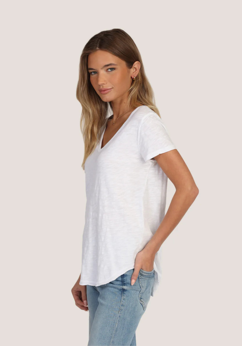 Side view of a woman wearing a white, V-neck t-shirt and blue jeans on a plain background