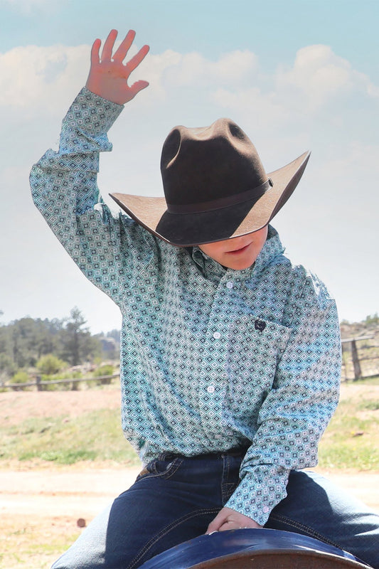 Boy wearing a turquoise patterned, Cinch shirt and cowboy hat outdoors with a clear sky.