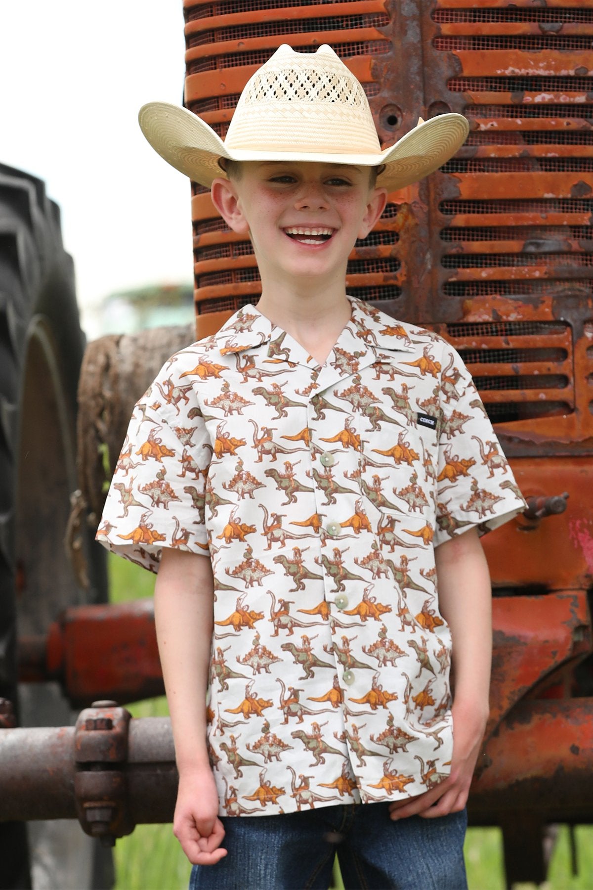 Young boy in front of tractor wearing cowboy hat and white short-sleeve snap shirt featuring cowboys riding dinosaurs