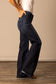 Side view of a woman wearing high waisted and dark washed Kimes Olivia flared jeans and a white t-shirt against a beige wall.