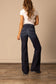 Back view of a woman wearing high waisted and dark washed Kimes Olivia flared jeans and a white t-shirt against a beige wall.