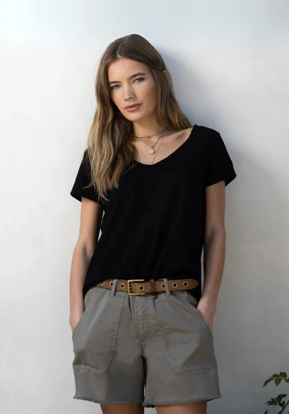 Woman wearing a black V-neck t-shirt and beige shorts with a brown belt against a white wall.