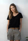 Woman wearing a black V-neck t-shirt and beige shorts with a brown belt against a white wall.