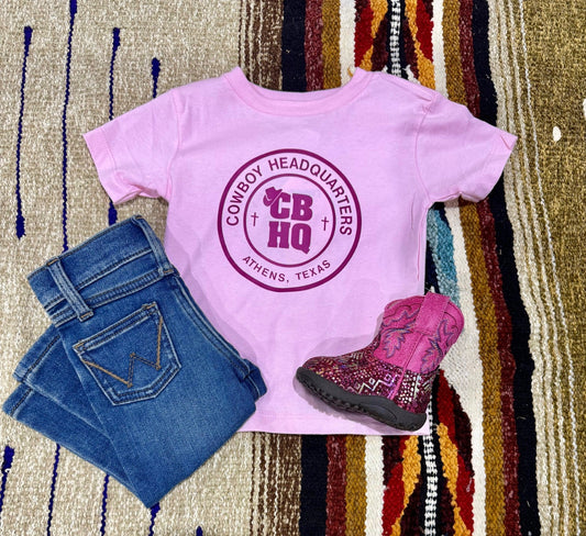 Pink t-shirt with 'Cowboy Headquarters' logo, blue jeans, and pink boots on a patterned rug.